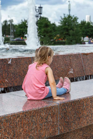 little girl sitting on the side of a fountain in a city park, back viewの写真素材