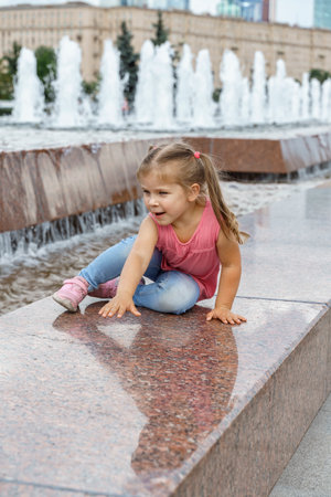 happy caucasian child girl crawling on the side of the fountain in the city parkの写真素材