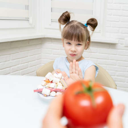 Cute little girl choosing between tomatoes and sweets, child rejects the vegetable and chooses candyの写真素材