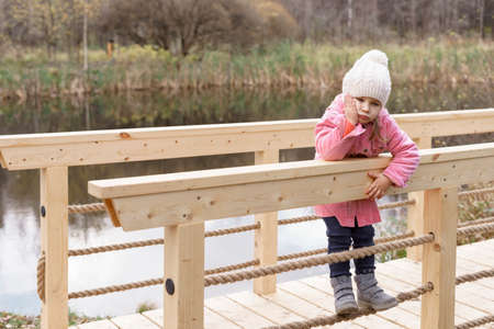 a sad little girl on a wooden bridge has a toothache and holds her cheek with her hand on an autumn dayの写真素材