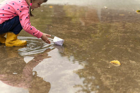 a little girl in a pink sweater and rubber boots launches a paper boat in a large puddleの写真素材