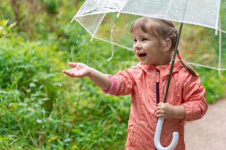 happy child walks in the park with a transparent umbrella on a rainy day. little girl laughs and catches dropsの写真素材