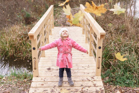 happy little caucasian girl in a pink coat in an autumn park on a wooden bridge throws yellow leaves upの写真素材