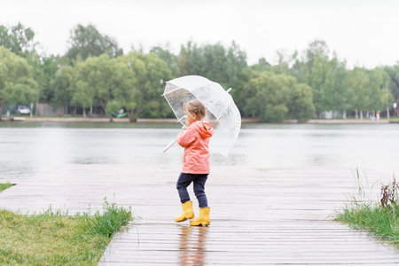 child girl in rainboots walks under an umbrella and looks at the raindrops on the pond.の写真素材