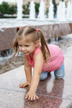 happy child girl crawling on the side of the fountain in the city park.の写真素材