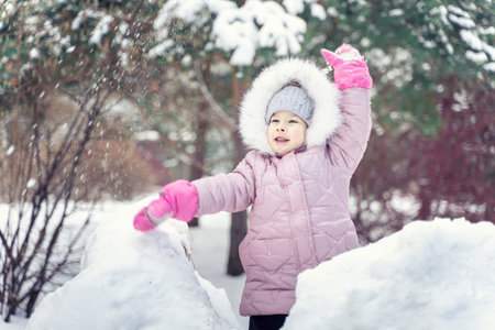 Smiling happy child girl in warm pink winter clothes throws a snowball in winter