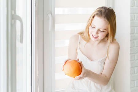 beautiful young woman in a white dress wants to peel a large grapefruit by the window in a white roomの写真素材