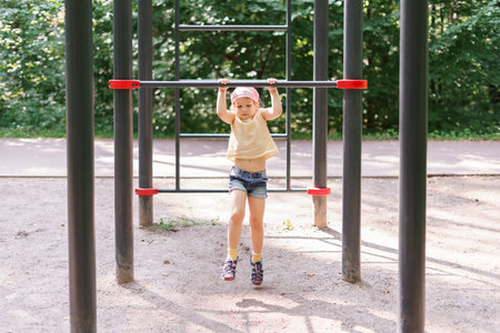 little girl pulls herself up on a horizontal bar in a city park on a summer dayの写真素材