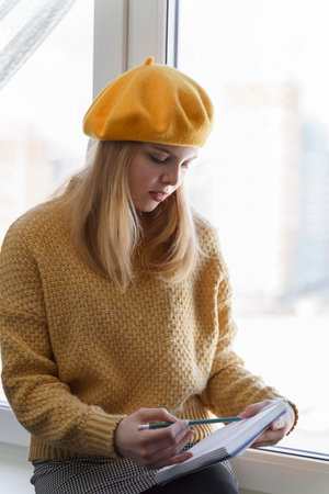 girl student in a yellow beret makes sketches in her sketchbook sitting by the window