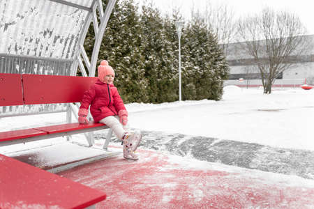 adorable kid girl with skates sits on a bench near the winter outdoor ice rinkの写真素材