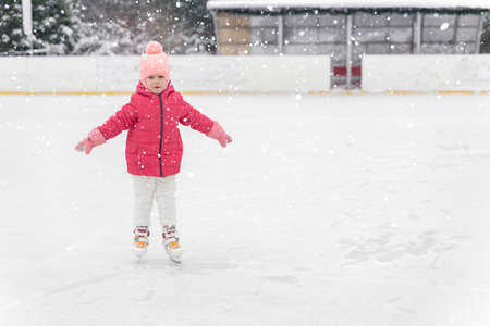 kid girl in a red jacket skates on a skating rink in heavy snow and blizzardの写真素材