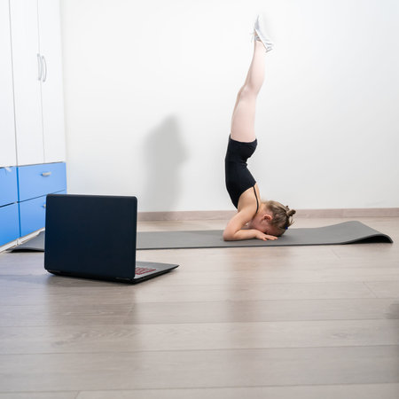 Fit nice aucasian little girl doing yoga and watching online lessons on laptop, exercising in the...