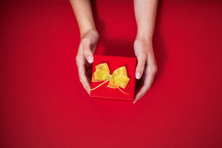 Christmas birthday red gift with a gold bow in the woman hands on the red backgroundの写真素材