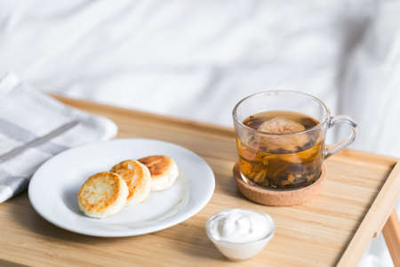 Yellow cottage cheese pancakes on the bed tray table with black tea and sour cream and napkin in the morningの写真素材