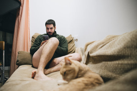 Man with a beard sitting on the sofa with a phone at home blurred catの写真素材