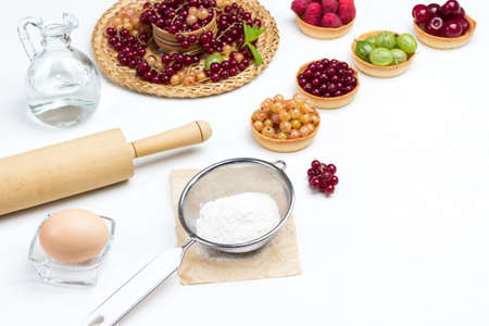 Flour in sieve, rolling pin and egg in glass small bowl. Decanter with water. Tartlets with berries. White background. Top viewの写真素材