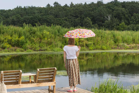 Woman with umbrella stands on bank of the river. Forest on the other sideの写真素材