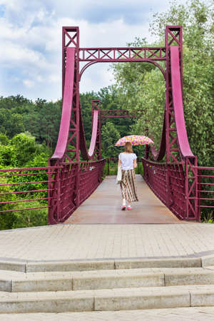 Woman with an umbrella is walking along the red iron bridge. Dense forest and blue sky.の写真素材