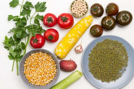 Chickpea, corn and mung bean grains in plates. Swing of yellow corn, tomatoes and parsley sprigs on table. White background. Flat layの写真素材