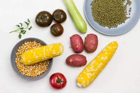 Swing of corn and corn grains in gray bowl. Tomatoes, potatoes and corn on table. Mash grains on plate. White background. Flat layの写真素材