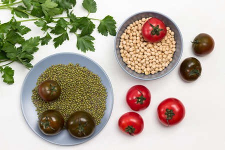 Chickpea grains and mung bean in gray plate. Tomatoes and sprigs of parsley on table. White background. Flat layの写真素材