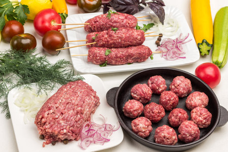 Raw minced meat on plate. Raw meat balls in frying pan. Raw kebab on wooden skewer. Vegetables and greens on table. White background.の写真素材