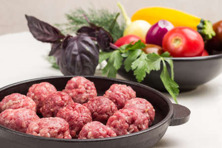 Raw meat balls in frying pan. Vegetables and greens in plate. Close up. White background.の写真素材