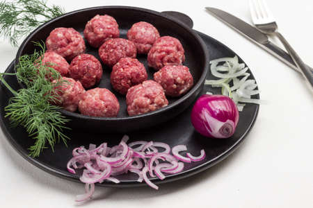 Raw meat balls in frying pan. Chopped onion on plate. Dill sprig, knife and fork on table. White background.の写真素材