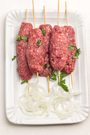 Raw minced meat on wooden skewers and chopped onion rings on white plate. White background. Flat layの写真素材