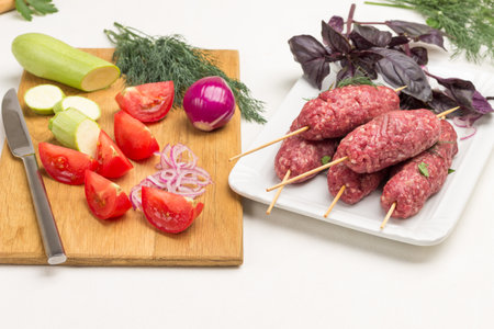 Sliced tomatoes and zucchini on cutting board. Raw minced meat on wooden skewers. Kebab cooking. White background.の写真素材