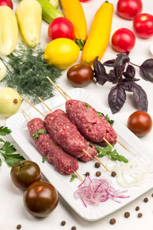 Raw minced meat on wooden skewers. Tomatoes, zucchini, peppers, dill and sprig of basil on table. White background.の写真素材
