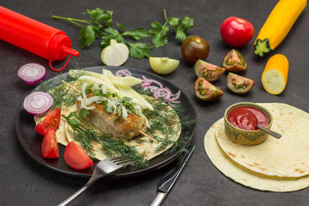 Meat kebab with vegetables and herbs on plate. Pita and chopped tomatoes on table. Black background. Top viewの写真素材