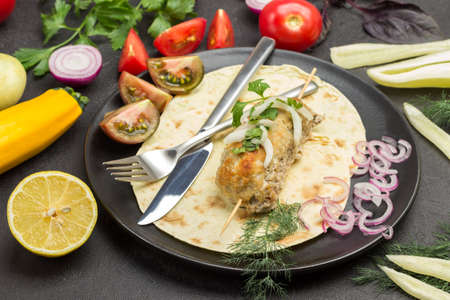 Kebab on skewer with tomatoes and onions on pita in plate. Knife and fork. Vegetables on table. Black background. Top viewの写真素材