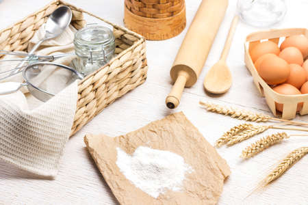 Flour on paper. Spikelets of wheat. Eggs in wicker box and spikelets of wheat. Glass jar, sieve and spoon in wicker box. White background. Top viewの写真素材