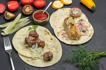 Kebab on skewer and meatballs with tomatoes and onions on pita. Tomato sauce in bowl. Vegetables on table. Black background. Top viewの写真素材