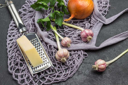 Mesh fabric bag with groceries. Garlic, onion and cheese on grater, sprig of parsley. Black background. Top viewの写真素材