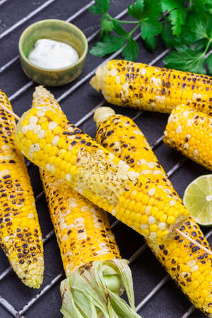Grilled corn cob on metal grill grate. Sauce in bowl and fresh parsley on table. Black background. Top viewの写真素材