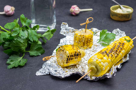 Grilled corn on foil. Fresh parsley on table. Sauce in bowl. Bottled water. Black background. Top viewの写真素材