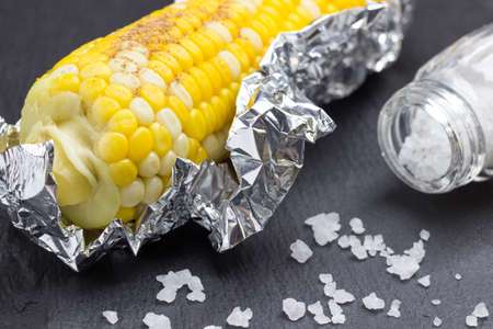 Boiled corn in foil. Salt shaker and salt on table. Black background. Close upの写真素材