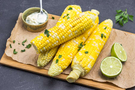 Boiled corn on wooden board. Lime and sauce in bowl. Parsley leaves. Black background. Top viewの写真素材