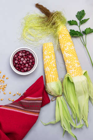 Cobs of raw corn with leaves and corn silk. Frozen berries in bowl. Red napkin.Gray background. Flat layの写真素材