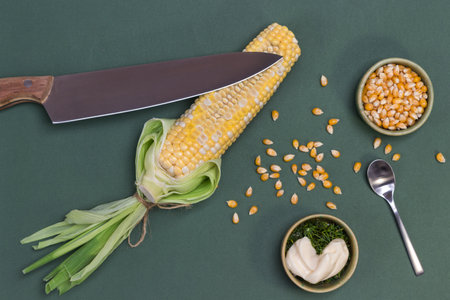 Knife on raw corn. Corn grains on table and in bowl. Sauce in bowl. Gray background. Flat layの写真素材