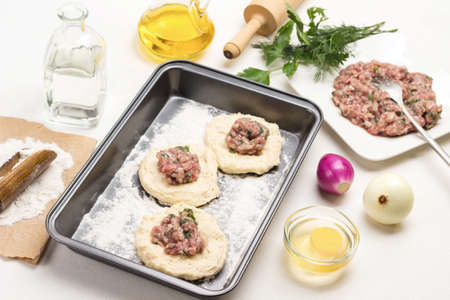 Filling of meat on pieces of raw dough on baking pan. Minced meat, fork and parsley sprigs on plate. White background. Top viewの写真素材