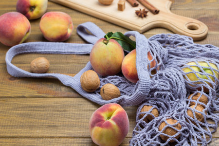 Peaches with green leaves in fabric mesh bag. Walnuts, cinnamon sticks and star anise on board. Wooden background. Top viewの写真素材