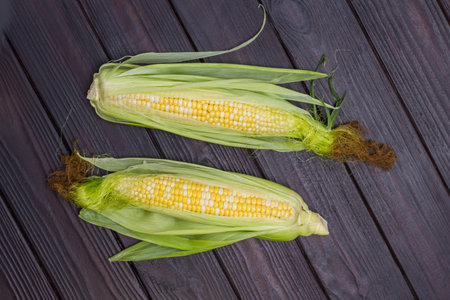 Two ears of corn with leaves and corn silk. Dark wooden background. Flat lay