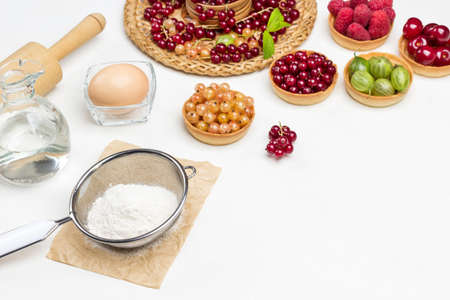 Flour in sieve, egg and rolling pin, bottle of water. Tartlets with berries. Red currants on wicker plate. Copy space. White background. Top viewの写真素材
