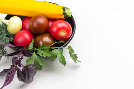 Set of vegetables and sprigs of basil and parsley in black bowl. Yellow zucchini and red tomatoes. White background. Copy space. Top viewの写真素材