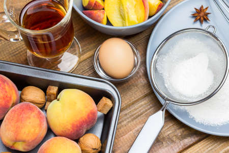 Ripe peaches in metal tray. Flour and sieve on gray plate. Glass of tea and sliced apples in gray plate. Wooden background. Top viewの写真素材