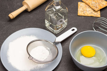 Flour and sieve on gray plate. Egg yolk in bowl. Decanter with water and cookies on table. Cooking. Black background. Top viewの写真素材