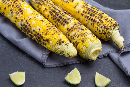 Three cobs of grilled corn on gray napkin. Lime slices on the table. Black background.の写真素材
